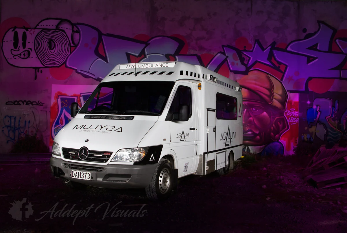 The original Asylum tattoo removal business: a converted white ambulance branded Asylumbulance, parked at night in front of a graffiti wall
